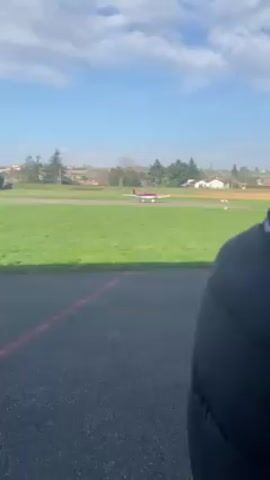 The video shows a group of people filming a small plane on a runway. The cameraman is setting up a large camera on a gimbal mount. The plane is red and white. The sky is blue with white clouds.