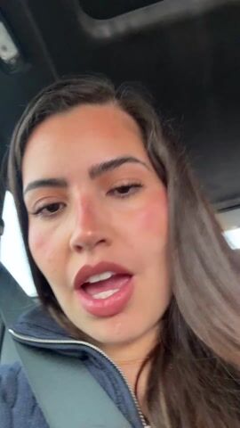 A person with long dark hair wearing a navy blue quarter-zip sweater and pearl earrings is recording a close-up video from inside a car. The camera angle is upward, capturing the person's face and a glimpse of the car's interior. The person is talking and moving their head slightly, with a concentrated focus on the camera. They have a notable tan or makeup blush on their face, particularly around their nose and cheeks. A seatbelt is visible across their chest.