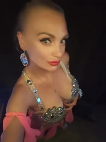 A woman with blonde hair pulled back, wearing a sparkly pink and gold belly dancing outfit, poses and smiles for the camera. She is also wearing large, dangling earrings and a thin necklace with a small pendant. The background is dark and out of focus.