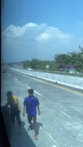 The video shows two young men by the side of a highway. One man, wearing a yellow shirt and a baseball cap, is hunched over a large tire, appearing to be inspecting or manipulating it. The other man, wearing a blue shirt and black shorts, is standing and looking in the same direction as the first man, with his left arm extended forward. The highway appears to be under a bright blue sky with scattered clouds. Trees and vegetation are visible on the right side of the road, with a white vehicle in the distance.
