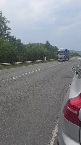 The video shows a road with a silver car in the foreground, and a large blue and silver semi-truck driving on the road. The truck is in the right lane and appears to be moving away from the viewer. The road is paved and has white lines marking the lanes. There are trees and foliage on either side of the road, suggesting a rural or natural environment. The sky is overcast.