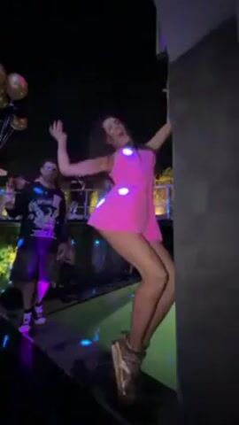The video shows a woman in a short, bright pink dress and platform sandals dancing at an outdoor party. She is holding onto a corner column for support and moving to the music. In the background, a man with a microphone appears to be performing or hosting the party. Other people are seen in the periphery, and there are balloons and a pool nearby. The lighting suggests it is nighttime.