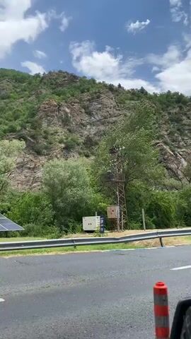 The video shows a roadside view with rocky, green-covered mountains in the background. The camera pans up to reveal the peaks of the mountains, which have little to no vegetation. In the foreground, there's a grey guardrail, a section of asphalt road, a generator, and a utility pole with equipment attached. The sky is partly cloudy.