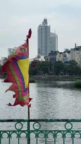 A tattered, colorful flag flutters on a pole in the foreground, with a body of water behind it. In the background, several buildings are visible, including a tall, modern skyscraper. The sky is overcast. The flag has stripes of red, pink, purple, yellow, and green.