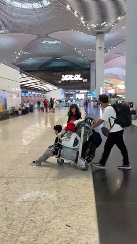 The video shows a family at an airport. A man wearing a white t-shirt and black pants pushes a luggage cart. A young girl with dark hair and a red shirt is sitting on the luggage cart. A young boy in a grey and white patterned outfit is also on the luggage cart. They are walking through a large, modern airport terminal with a high, curved ceiling and a polished floor. In the background, there are information desks, signs, and other travelers.