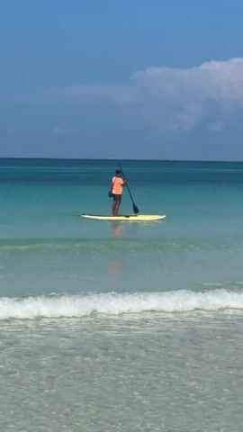 A person stands on a yellow paddleboard in clear turquoise water. They are wearing an orange shirt and dark shorts. The person is holding a paddle and appears to be paddling the board. A bag is slung over their shoulder. The ocean extends to the horizon under a clear blue sky with scattered white clouds. Gentle waves break on a sandy beach in the foreground.