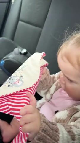 A baby is in a car seat. The baby is wearing a pink shirt and a patterned cardigan. The baby is holding a red and white striped blanket to its face. The baby's face is partially obscured by the blanket. The video shows the baby from the chest up. The background is the interior of a car, with black seats.