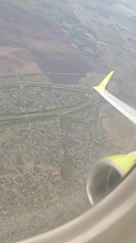 From the perspective of an airplane passenger, a view outside the window shows a landscape below. The scene captures a mix of agricultural fields, with patterns of dark and light soil suggesting different crops or land use. A dense cluster of buildings, resembling a small town or large residential area, sits prominently in the center of the frame, bisected by what appears to be a highway. The aircraft's wing, identifiable by its bright green tip, is visible in the foreground, along with a portion of the engine, offering a sense of scale and context to the aerial perspective. The overall image suggests a rural or suburban setting, viewed from a relatively low altitude during daylight.