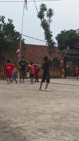 A group of children are gathered outside a building that appears to be a school, indicated by the sign that reads "SON JAMARAGA". The children are positioned in a courtyard-like area with a concrete surface. Some of the children are wearing red shirts, while others are wearing black shirts. They appear to be participating in some kind of activity, possibly a game or a performance, as some of them are holding Indonesian flags and making coordinated movements. The weather appears to be overcast. There are trees and a building with a red roof in the background. One child in a black shirt and shorts is walking towards the right of the frame, away from the camera, while another child in a black shirt is facing the camera and holding a flag.