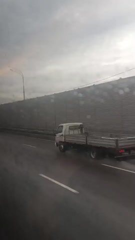 The video shows a highway scene under a cloudy sky. The road is wet, suggesting recent or ongoing rain. A metal sound barrier runs along the right side of the highway, with a corrugated texture and circular patterns on its surface. Several vehicles are visible on the road, including a white truck on the right, a silver car and a dark blue car on the left. The cars are moving, indicating traffic on the highway. The perspective appears to be from inside another moving vehicle, looking out the window. There are water droplets on the window, further suggesting rain or wet conditions. The overall impression is a gloomy, overcast day with traffic moving along a wet highway.