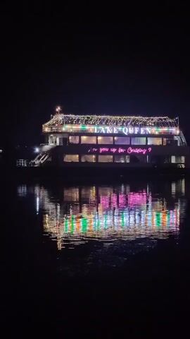 A nighttime shot captures a boat named Lake Queen illuminated by decorative lights. The lights spell out the boat's name on the upper deck and also display the text "I'm you up for cruising?". Colorful lights adorn the side of the boat and are reflected in the dark water below.