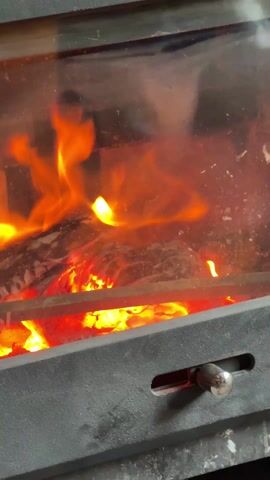 A close-up video shows a fireplace with a glass door. Inside, a fire burns brightly, with flames flickering around pieces of wood and glowing embers visible below. The frame of the fireplace is dark gray, and a silver handle is visible on the lower right side. The glass door is slightly dirty, and an indistinct reflection can be seen.