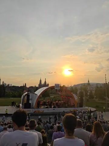 The video shows an outdoor concert at sunset. A band is performing on a stage with a large, arched backdrop. The audience is seated in tiered seating, facing the stage. The sun is setting behind the stage, casting a warm glow over the scene. In the background, there are several buildings with spires, possibly churches or government buildings. The sky is a mix of blue and orange, with some clouds. The overall atmosphere appears to be festive and relaxed.