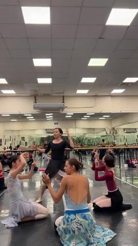 A group of young women are practicing ballet in a dance studio. There are five dancers in the foreground, all wearing ballet attire. One dancer, dressed in black, stands and instructs the others who are seated on the floor. The studio has mirrors on the wall and ballet barres.