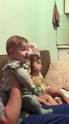 A low-angle, medium shot captures three young children sitting on a couch, looking towards the right side of the frame. In the foreground, a baby, held by an unseen adult, is positioned on the left. The baby has fair skin, light brown hair, and is wearing a grey long-sleeved shirt with a floral pattern and light green tights. The baby's head is turned to the right, with eyes wide and mouth slightly open, as if reacting to something. To the right of the baby, a slightly older child with dark brown hair and bangs is seated. This child is wearing a light blue shirt with a ruffle detail and appears to be smiling. Behind this child and further to the right, another child with dark hair and wearing a light-colored shirt with a cartoon character print is visible. This child also appears to be looking off-frame to the right. The couch is upholstered in a dark brown fabric, and a patterned pillow is visible behind the children. The background is a pale green wall, with a small brown cloth hanging on the upper right. The lighting is dim, suggesting an indoor setting, possibly an evening. The image is slightly grainy.
