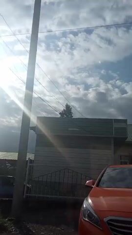 The video shows a power pole with cables stretching across the frame. Sunlight glints off the cables and pierces through a partly cloudy sky. In the foreground, a dark blue hatchback car is parked next to a building with a wooden exterior. The ground appears to be dirt or gravel. In the distance, there is vegetation and possibly rolling hills. The camera pans slightly to the right, revealing more of the sky and the power pole.