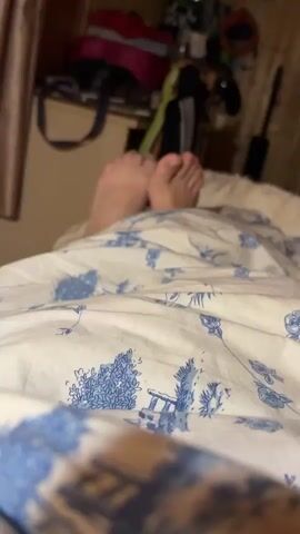 A close-up shot shows a pair of bare feet peeking out from under a white comforter patterned with blue and white chinoiserie designs. The feet are positioned in the center of the frame, with the toes pointing upwards. The background is blurred, but hints at a cluttered shelf with bags and other items. The lighting is dim, suggesting an indoor setting, possibly a bedroom.