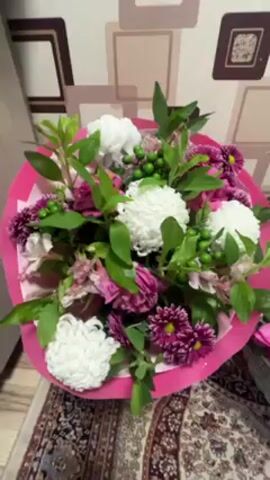 The video shows a bouquet of flowers on a carpet. The bouquet is primarily white and pink, with some green accents. The white flowers appear to be chrysanthemums. The pink wrapping contrasts with the brown patterned carpet. The background is a wall with square, framed patterns.