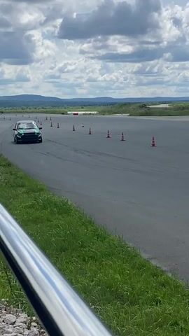 A black and green race car with the number 9 on its door drives on a paved track. Orange cones are placed along the side of the track. In the background, there are hills, a body of water, a large inflatable building, and several parked vehicles. The sky is cloudy.