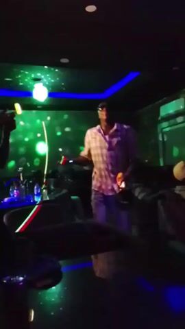 The video shows a person dancing in a dimly lit room with colorful lights. The person is wearing a plaid shirt and jeans. They are moving their arms and body to music. Other people are sitting in the background. The room has a dark ceiling with blue lighting accents and a decorative light fixture. The floor is dark and reflective. Other lighting includes vertical bars of alternating green and red lights.