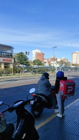 The video shows a street scene in a city. The camera is oriented vertically. In the foreground, two motorcycles are parked. One motorcycle has a red delivery bag with a white letter "P" logo on the back. A person wearing a blue helmet and a gray jacket is sitting on this motorcycle, facing away from the camera. Another person, also wearing a gray jacket, is seated on the second motorcycle in the foreground, facing towards the camera. Behind the motorcycles, there is a street with traffic. On the sidewalk, there is a trash can and a bus stop sign. In the background, there are buildings of varying heights, including some tall apartment buildings, and trees. The sky is clear and blue. There are traffic lights and streetlights visible. A digital display shows "CONO 1000".