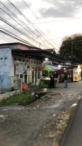 A street scene at dusk shows a building with a covered walkway in front. Underneath the walkway, several food stalls are set up. One stall has a green and yellow cart with an umbrella, where two people are standing, possibly vendors. Another person is standing near the cart. Further down the walkway, another food stall is visible with a yellow sign. A few people are seen sitting or standing near the stalls. The ground is paved, and there is some debris scattered around. Power lines are strung across the sky.