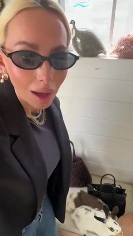 The video shows two women interacting with several rabbits in a small enclosure. The enclosure appears to be indoors, with artificial grass-like flooring and a wooden structure in the corner. The women are wearing casual clothing. One woman is wearing a black blazer and sunglasses. She is seen feeding the rabbits a green leafy substance from a bowl. The other woman is bent over, also interacting with the rabbits. Several handbags are on the floor near the enclosure. The woman wearing the blazer looks at the camera and smiles.