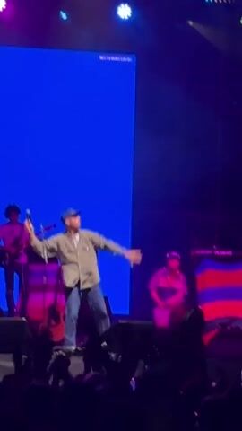 A man wearing a light-colored collared shirt, blue jeans, and a baseball cap stands on a stage in front of a large blue screen. He is dancing and gesturing with his arms. To his left, a person with curly hair plays a guitar. To his right, another person is kneeling. In the foreground, the hands of audience members are visible, many holding up phones to record the performance. The stage is lit with pink and purple lights.
