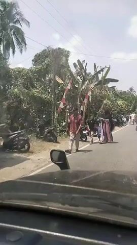 The video shows a road scene with people and motorcycles. On the left side of the road, there are many parked motorcycles lined up. Behind the motorcycles, there are banana trees and other vegetation, with some red and white flags visible. Several people are walking on the side of the road and on the road itself. A man in a checkered shirt and a cap is walking on the left side of the road. Further down the road, a group of people are walking together. On the right side of the road, more motorcycles are parked, and traffic appears to be moving. The video is filmed from inside a vehicle, with the dashboard and a side mirror visible in the foreground. The weather appears to be sunny with some clouds.
