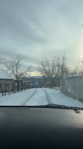 The view is from the interior of a vehicle looking out the windshield. It is a winter scene with snow on the ground and along the sides of a narrow road. Tire tracks are visible in the snow. The road slopes uphill slightly. There are bare trees on either side of the road and fences line the properties. In the distance, there is a vague, mountainous or hilly landscape visible. The sky is overcast with the sun appearing as a bright spot behind the clouds.