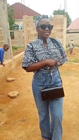 A woman with dark skin is standing in an area that appears to be under construction. She is wearing sunglasses, a patterned black and white shirt, and high-waisted blue jeans. She has a head wrap that matches her shirt. She is carrying a small black purse with a reptile-like texture. Behind her, there is a partially constructed brick building. In the background, there are other people and buildings with red roofs. The ground is dirt. The woman is smiling and holding her purse.