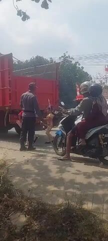 A red truck is parked on the right side of a paved road. To the left of the truck, a person wearing a red headscarf and a white shirt is standing. Next to them is a person in a grey uniform and a cap. In front of the truck, on the road, a blue and white motorcycle is stopped. A person wearing a helmet is driving the motorcycle, and a person wearing a hijab and a maroon shirt is sitting behind them. Further to the right, a white car is stopped behind the motorcycle. Another motorcycle is partially visible on the far right. The sky is overcast.