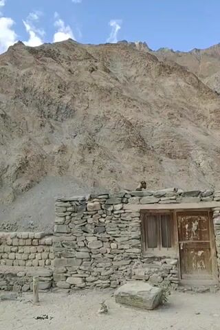 The video pans across a barren, mountainous landscape. In the foreground, a small, rustic stone building with a wooden door and window is visible. Behind it, a larger, more modern structure made of concrete and plaster is seen, with several doorways and windows. The terrain is dry and rocky, with sparse vegetation. The sky is blue with a few scattered clouds.