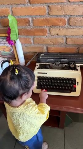 A young child with dark hair and a yellow shirt is standing at a wooden table and touching the keys of an old-fashioned typewriter. The typewriter is white with black keys and red accents. Behind the typewriter is a brick wall. To the left of the typewriter, there is a white bottle with a green leaf and pink flowers sticking out of it.