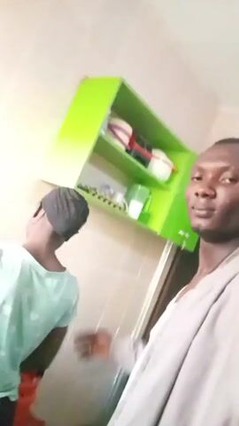 The video shows two individuals indoors. A man, dressed in a light grey jacket over a white shirt, stands behind a younger woman. The woman is wearing a green shirt with a black head covering. The man places his arms around her, hugging her tightly. The setting appears to be a kitchen or a room with kitchen cabinets, which are green. The walls are light colored, possibly white or off-white. There is a sound of banging in the background.