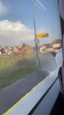 The video shows a view from inside a moving train. The camera is focused on the window, revealing a roadside scene with houses and utility poles passing by. The sky is partly cloudy. A person can be seen sitting in the train seat.