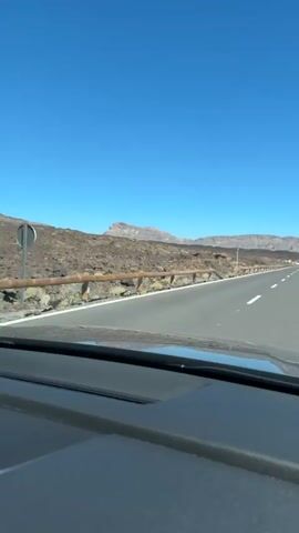 The video is a first-person perspective from inside a car traveling on a paved road. The road is asphalt with white lane markings and is bordered by a wooden guardrail on the right. To the left of the road, there is a rugged, rocky, and barren landscape with some sparse vegetation. In the distance, there are rolling hills and mountains under a clear blue sky. The dashboard of the car is partially visible in the foreground. The car appears to be moving at a steady pace.