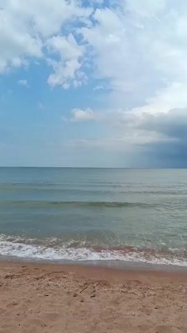 The video shows a beach with calm ocean waves. The sky is cloudy and appears to be preparing for a storm, with dark clouds gathering in the distance. The sand on the beach is light brown. The overall scene is serene despite the impending weather change.