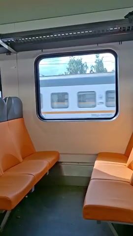 The video shows the interior of a train car with two rows of orange seats facing each other. A window on the right side of the frame shows another train passing by outside. The passing train is white with a yellow stripe. Trees are visible in the background outside the window. The interior of the train car is clean and modern. The lighting suggests it is daytime.