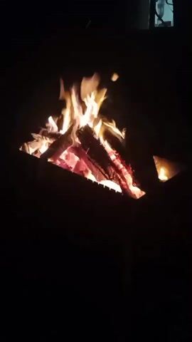 The video shows a close-up shot of a bonfire at night. The fire is burning brightly with flames reaching upwards. Sparks are visible rising from the fire. The logs in the fire are red hot. The background is dark and out of focus, with a faint hint of a structure in the upper right corner. The camera is stationary.