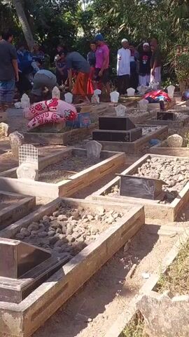 A group of people are gathered in a cemetery. The graves are marked with stones and some have headstones. Some of the people are dressed in traditional religious attire. Some are standing and observing, while others appear to be tending to the graves. One person is bent over a grave, possibly placing something on it. The environment appears to be outdoors with trees and vegetation visible in the background. The lighting suggests it is daytime.