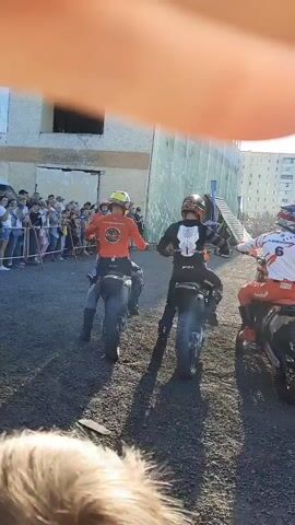 The video shows four motorcycle riders lined up on dirt bikes. They are all wearing protective gear, including helmets, and racing suits. The riders are facing away from the camera, and the camera is focused on their backs and the motorcycles. The environment appears to be an outdoor track or arena, with spectators in the background and buildings behind them. The riders are positioned in a staggered formation, with the rider on the far left wearing a red suit, the next rider wearing a black suit, the third rider wearing a white and orange suit with the number 6, and the rider on the far right wearing a white and orange suit with the number 150. The video captures the moment before the race begins, with the riders waiting for the signal to start.