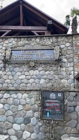 The video shows the exterior of a Russian Orthodox monastery, the "Monastyr Krestovaya Pustyn," which translates to "Monastery of the Cross Hermitage." The camera pans upward from a stone wall with signage and a set of rules for entering the monastery. The signage on the wall reads “Monastyr Krestovaya Pustyn” in Russian. The wall is constructed from a variety of light and dark grey rounded stones with some red bricks. As the camera pans up, the architecture of the monastery is revealed, including stone arches, rooftops, and gilded onion domes with crosses atop them. A black metal staircase leads to the entrance, which includes large wooden gates. The sky above is blue with white clouds.