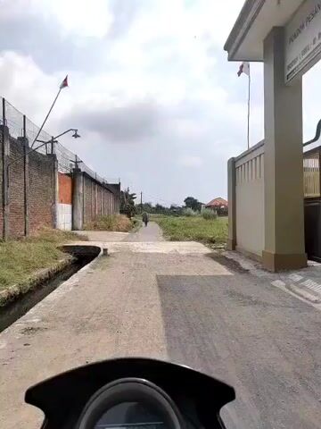 The video, shot from the perspective of a motorcycle rider, shows a narrow paved road leading into a rural area. A person on foot is walking away from the camera on the road. To the left, there is a brick wall with a chain-link fence on top, and tall dry grass. To the right, there is a beige building with a white trim and a small red and white flag flying. The road is bordered by overgrown green grass and some scattered bushes. In the distance, there is a house with a reddish-orange roof. The sky is cloudy with some patches of blue. The motorcycle's dashboard is partially visible in the foreground, displaying the number 14.