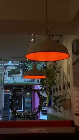 The video shows the interior of a restaurant or cafe, decorated with festive elements. Two large, off-white industrial-style pendant lights with orange interiors dominate the frame. In the background, a window display features illuminated deer figures and a large wreath adorned with fairy lights. A person in a dark coat and beanie walks past the window. The interior decor includes hanging plants and a rainbow flag, adding a touch of color. The focus remains on the lamps and their ambient glow, with the exterior scene providing a blurry, atmospheric backdrop.