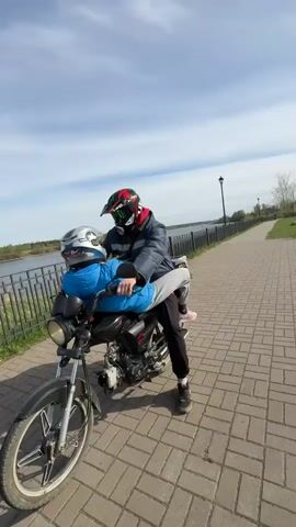 A man wearing a full face helmet and jacket sits on a motorcycle on a brick paved walkway. In front of him, a younger person with a helmet and jacket is sitting on the gas tank, facing the man. The motorcycle is angled slightly toward the viewer. In the background is a body of water with a metal fence and a grassy bank beyond it. To the right is a sidewalk running parallel to the water, along with a lamppost and some trees in the distance. The sky is partly cloudy. The motorcycle engine is revving.