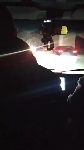 The video, shot from inside a moving car, shows the view through the windshield at night. The headlights of the car illuminate the road ahead, with another car visible in the distance, its red taillights prominent. The interior of the car is dimly lit, revealing two occupants. The driver is visible from the back, and their hands are on the steering wheel, while a passenger is seen to the side, wearing a hooded jacket. The dashboard and rearview mirror are visible. The overall setting conveys a nighttime drive, with limited visibility due to darkness.