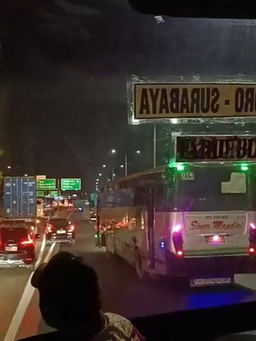 The video shows a nighttime view from inside a vehicle, likely a bus, looking out at a busy highway. Traffic is heavy, with many cars and trucks visible. The road is illuminated by streetlights and vehicle headlights. A large bus with "Sinar Mandiri" written on the back is in the foreground, to the right. There are signs with directional information, including one that appears to say "SURABAYA" and "Sidoarjo." The overall impression is one of a congested commute or journey on a highway at night.