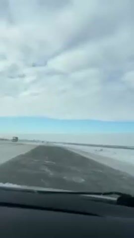 The video depicts a drive on a highway in snowy conditions. The sky is cloudy with patches of blue. Snow is visible on the shoulders of the road. The road surface is damp. A semi-truck can be seen in the distance. The video is shot from inside a vehicle.