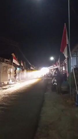 The video shows a nighttime street scene with Indonesian flags displayed on poles. A bright light source, possibly headlights from an approaching vehicle, illuminates the street in the distance, creating a streak of light. Several people are visible on the sidewalks and on motorcycles. The street is lined with buildings that appear to be shops or stalls. The overall atmosphere is that of a bustling street at night, possibly during a festival or event, given the flags and the bright lights.
